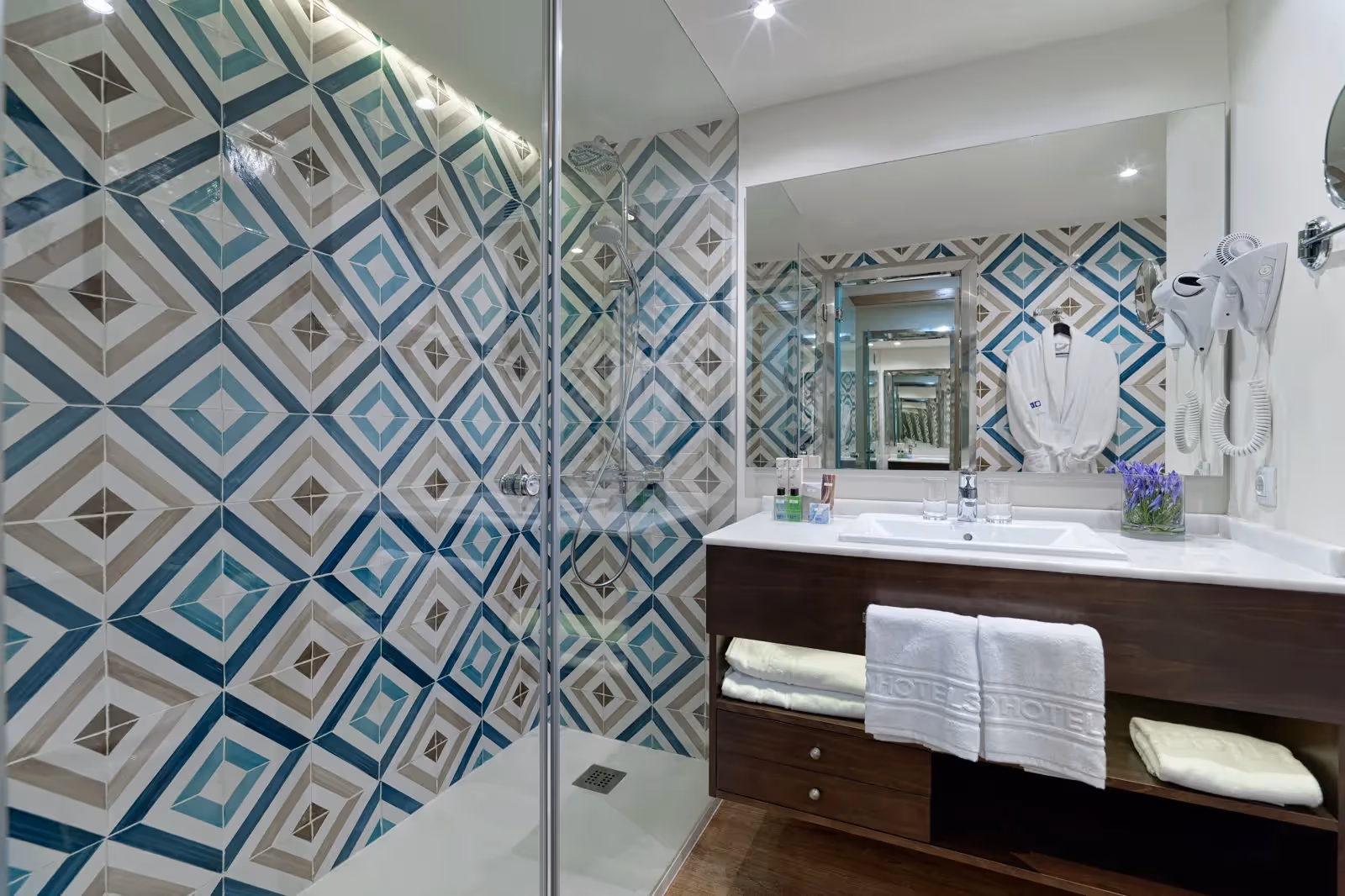 Badkamer met een inloopdouche, wandtegels met een blauwgrijs ruitpatroon, een wastafel en een badjas bij H10 Conquistador in Playa de las Américas