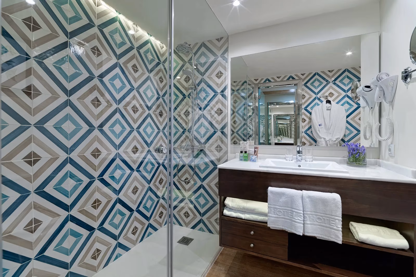 Badkamer met een inloopdouche, wandtegels met een blauwgrijs ruitpatroon, een wastafel en een badjas bij H10 Conquistador in Playa de las Américas