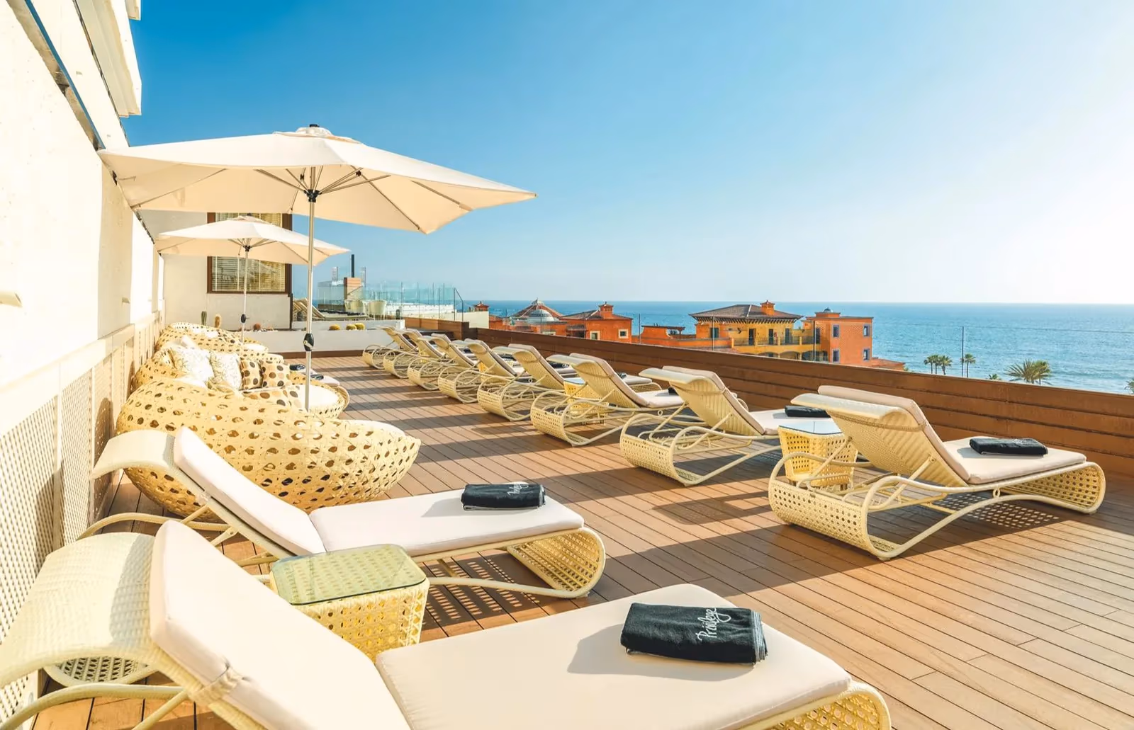 Dakterras met rieten ligstoelen met zwarte Privilege-handdoeken en parasols, met uitzicht op de kust bij H10 Conquistador in Playa de las Américas