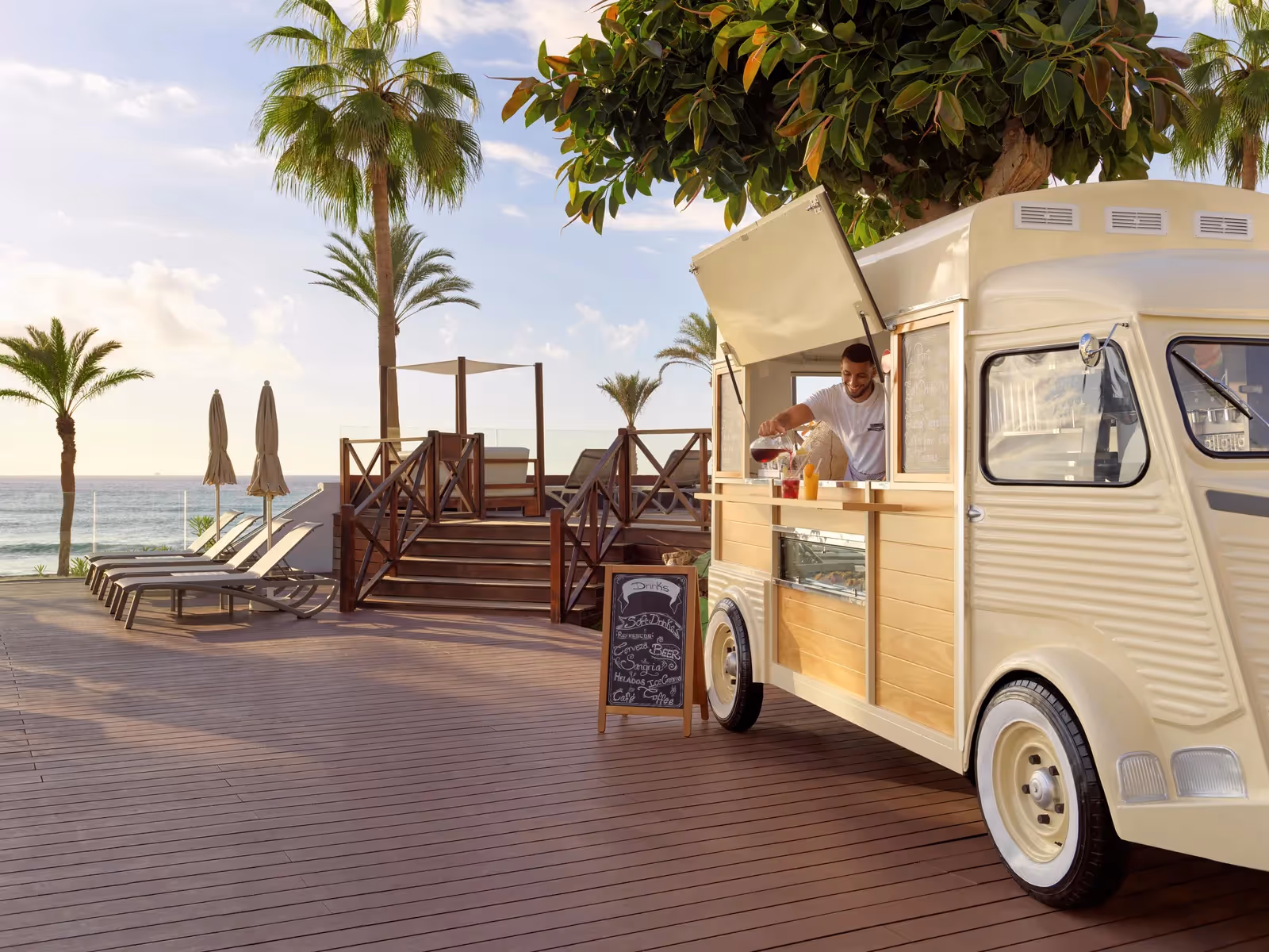 Crèmekleurige foodtruck op een houten terras aan de zee, met ligbedden en palmen bij H10 Conquistador in Playa de las Américas