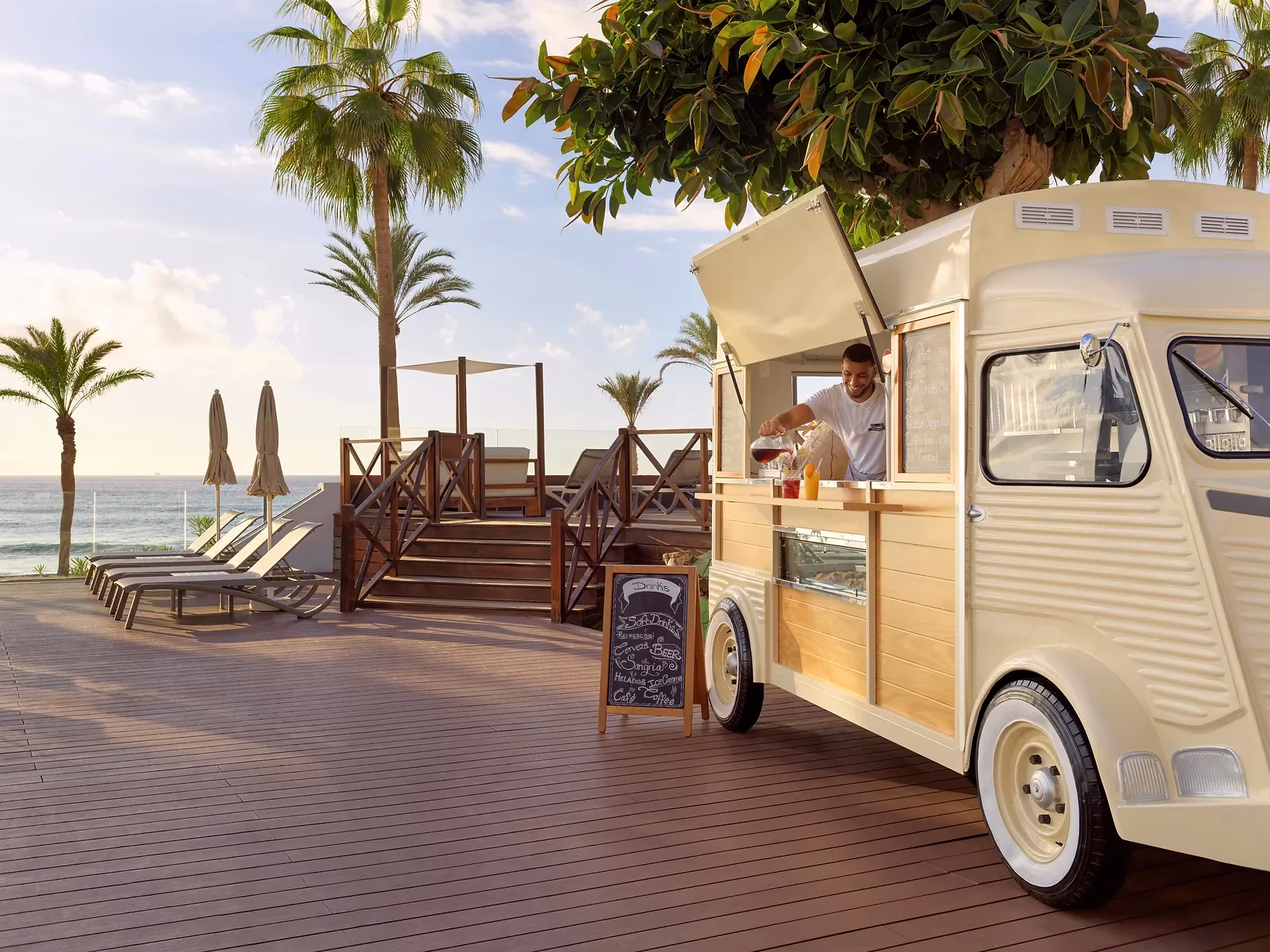 Crèmekleurige foodtruck op een houten terras aan de zee, met ligbedden en palmen bij H10 Conquistador in Playa de las Américas