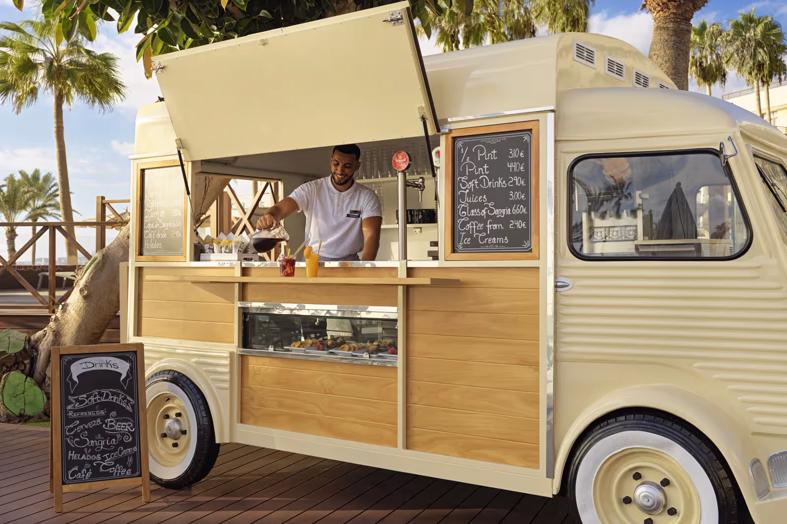 Crèmekleurige foodtruck met een medewerker die drankjes inschenkt en krijtborden met het drankaanbod bij H10 Conquistador in Playa de las Américas