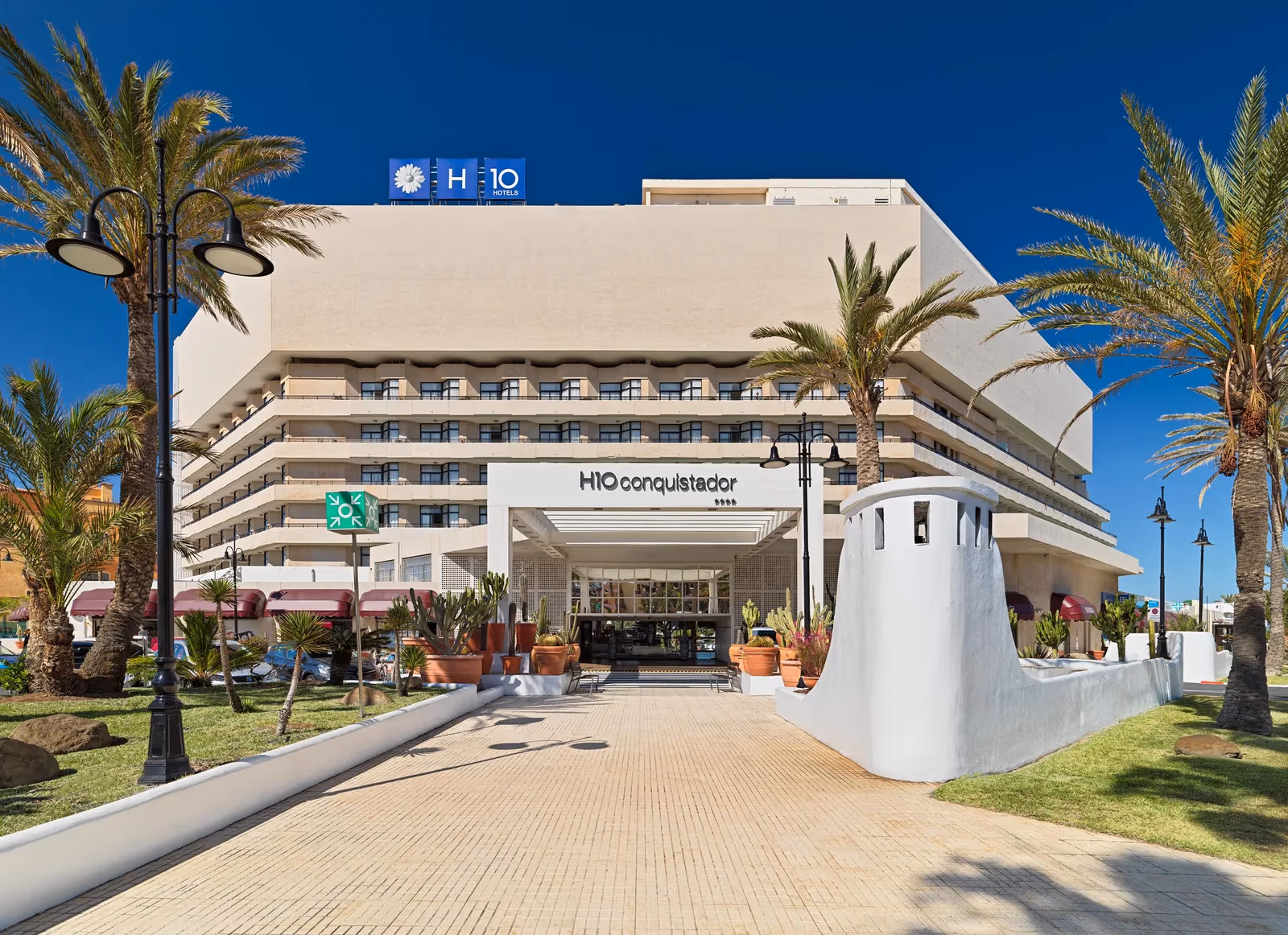 Hoofdingang met het H10 Conquistador-bord boven een overkapte entree, een tegelpad en palmen bij H10 Conquistador in Playa de las Américas