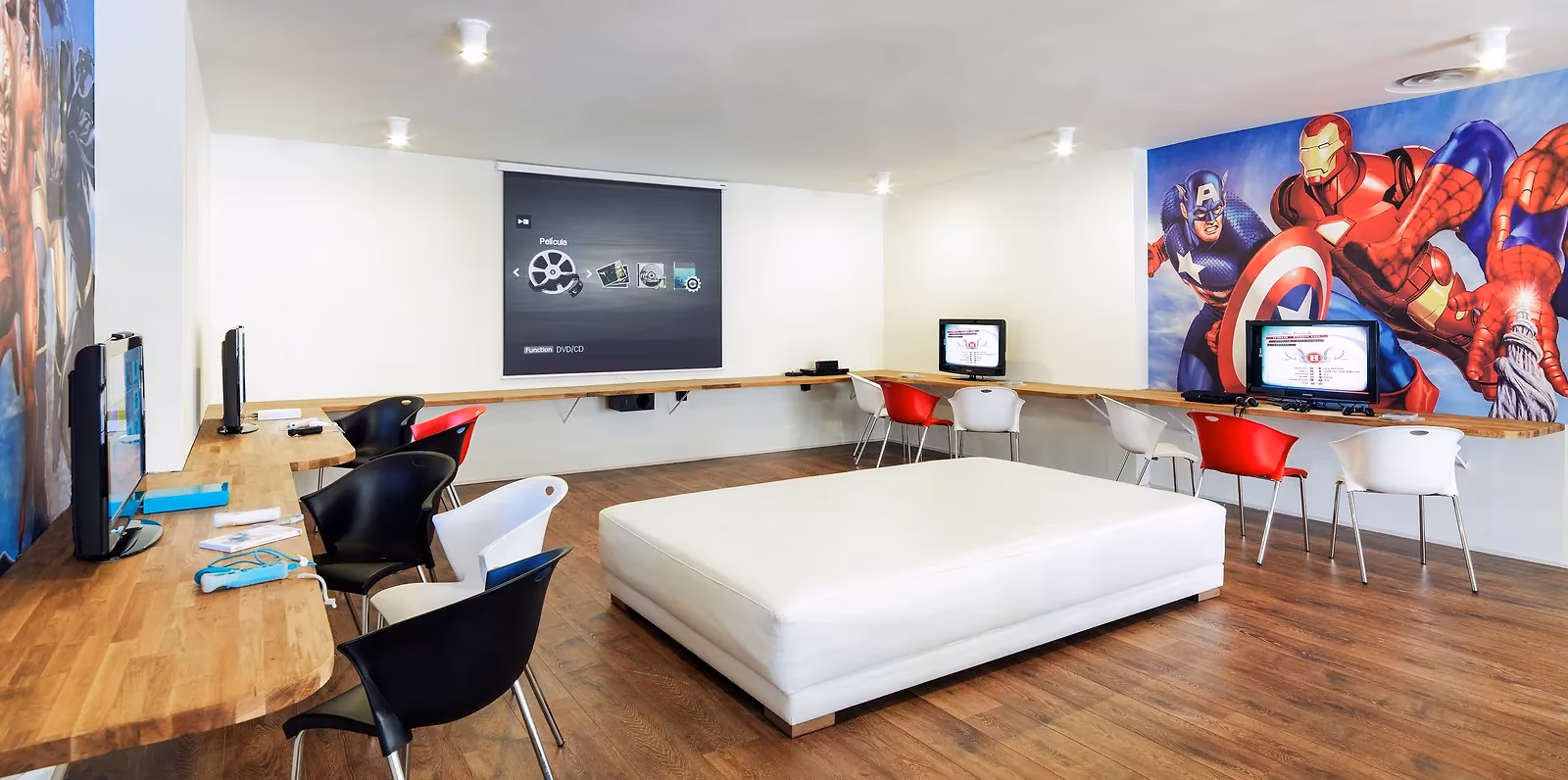 Ruimte met spelconsoles op schermen, een filmprojector, rode en zwarte stoelen, een grote witte poef en een muurschildering van superhelden bij H10 Conquistador in Playa de las Américas