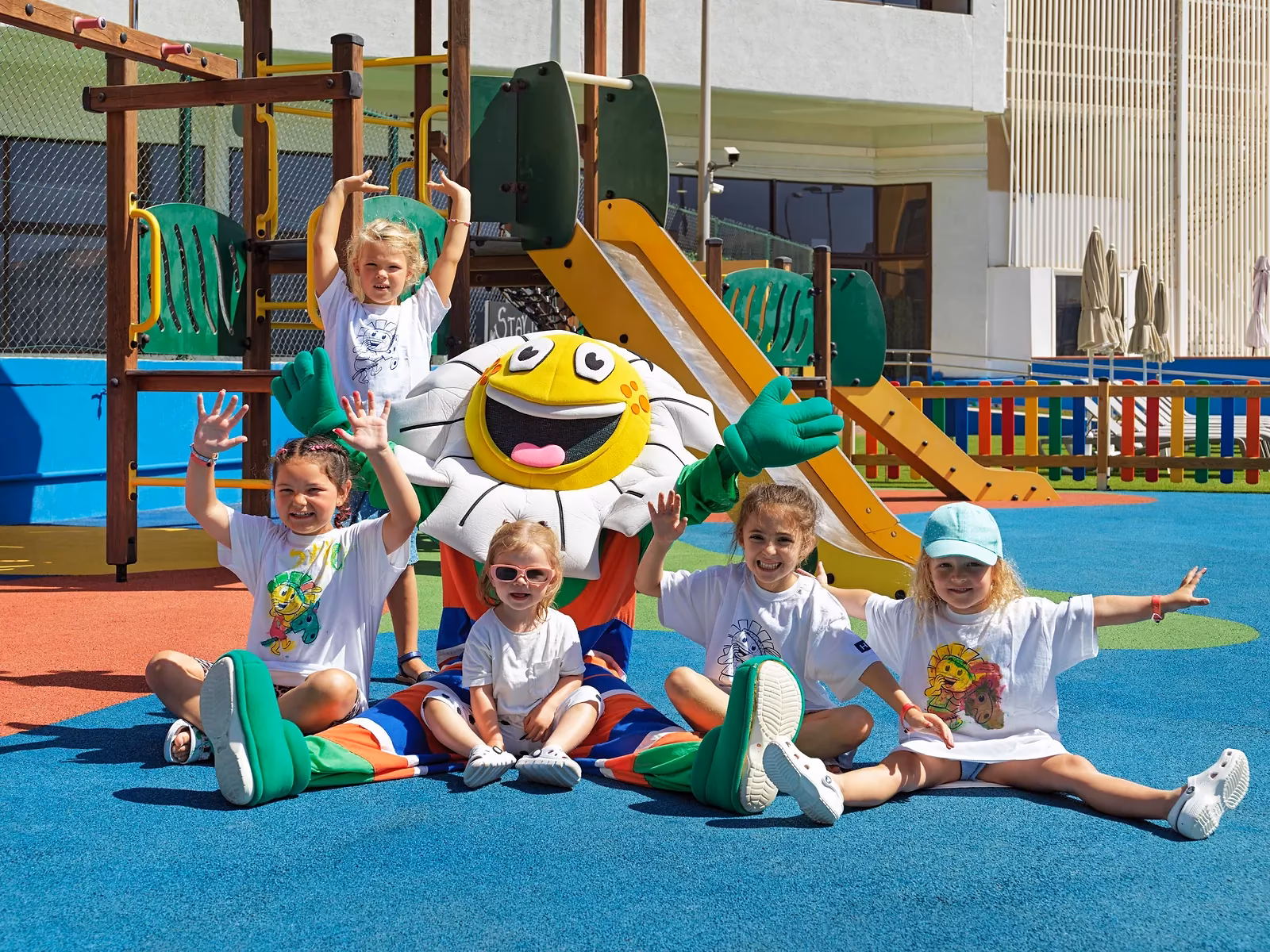 Vier kinderen in gelijke T-shirts bij een madeliefjesmascotte op een blauwe speelvloer bij H10 Conquistador in Playa de las Américas