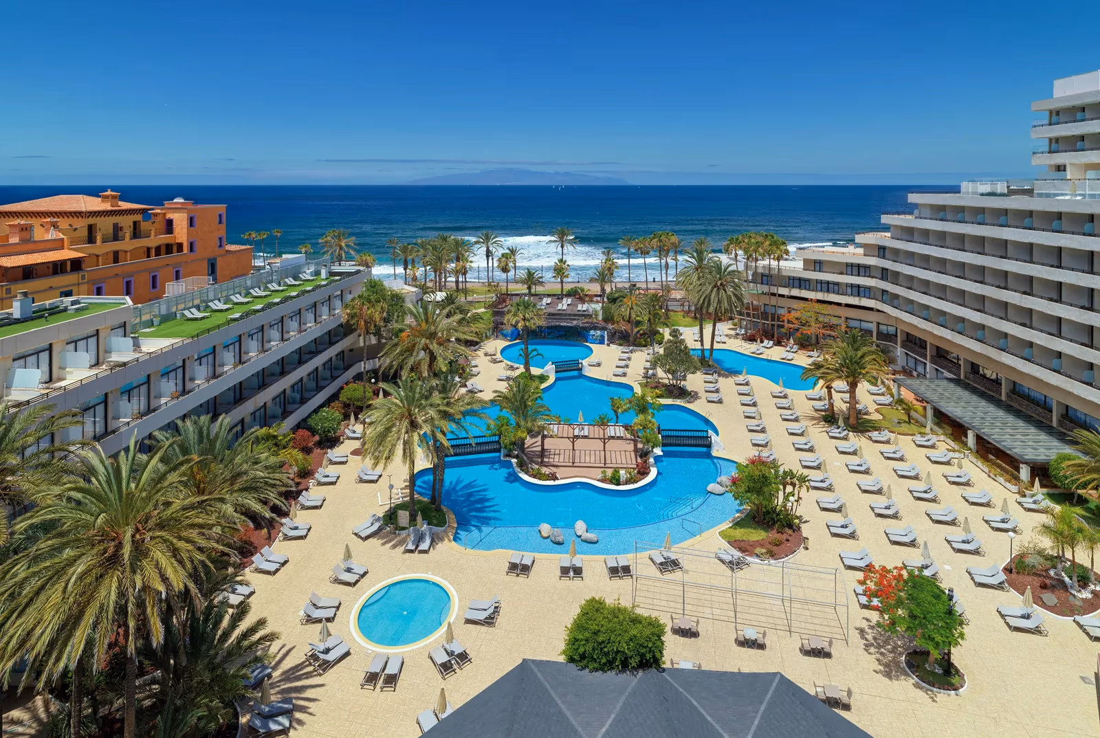 Luchtfoto van het hotelterrein met de zwembaden, de palmentuin en de oceaan aan de horizon bij H10 Conquistador in Playa de las Américas