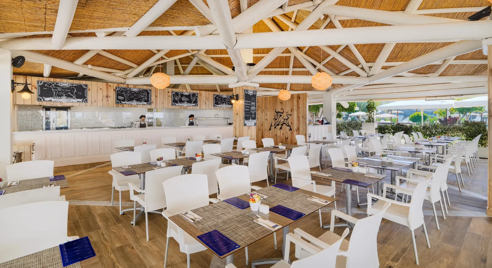Overdekt restaurant met witte stoelen, houten tafels, een rieten dak en krijtborden met vleessoorten bij H10 Conquistador in Playa de las Américas
