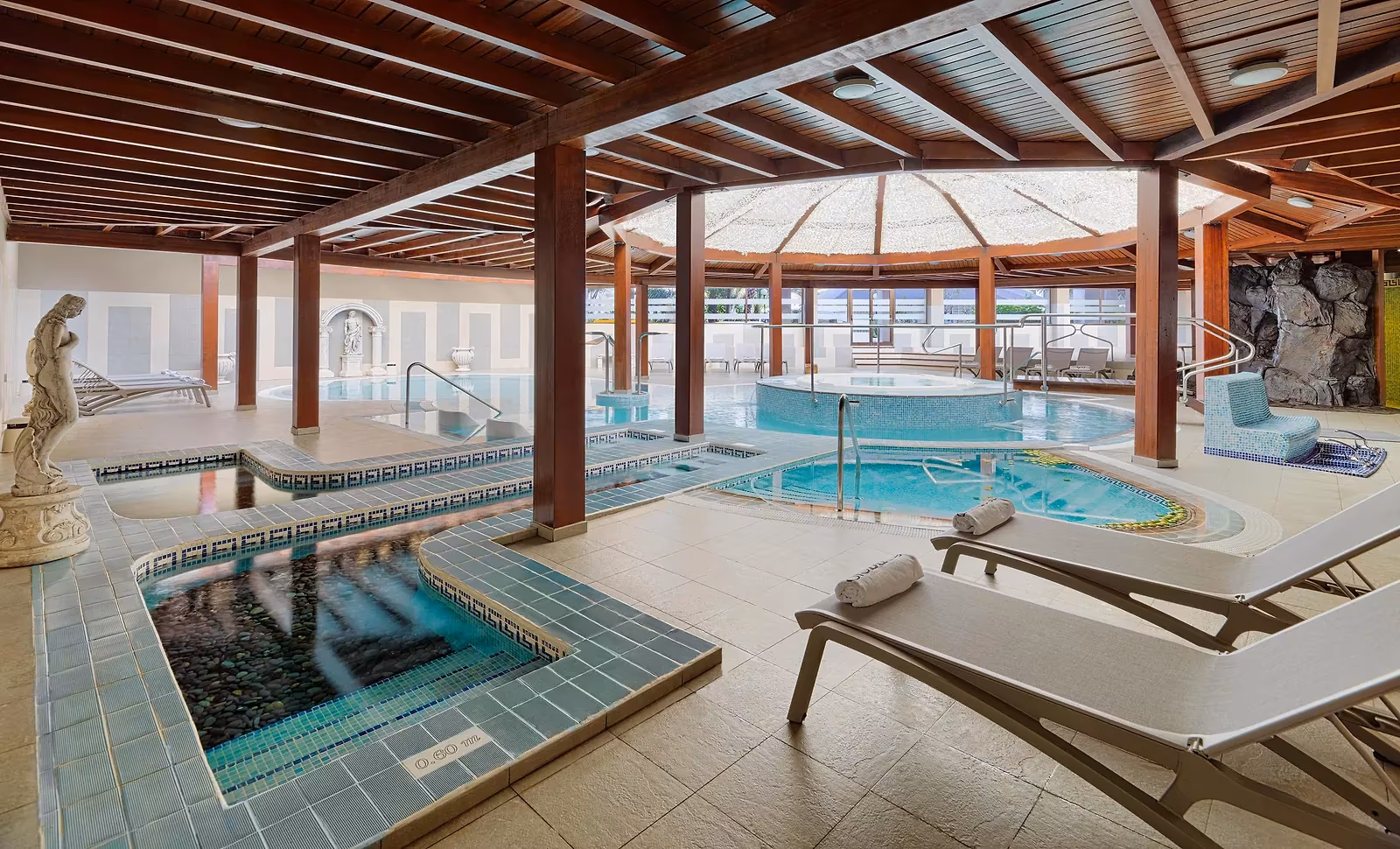 Overdekt wellnessparcours met meerdere baden, een ronde jacuzzi en ligbedden met opgerolde handdoeken onder een houten dak bij H10 Conquistador in Playa de las Américas