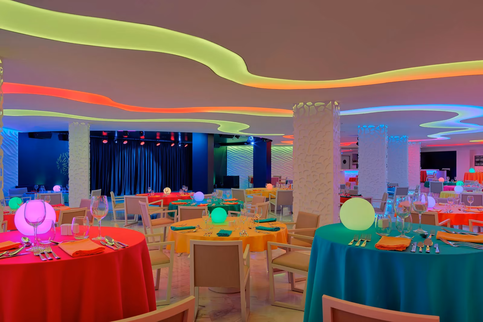 Feestelijk verlichte zaal met ronde tafels in felle kleuren, lichtgevende bollen en een podium met blauw gordijn bij H10 Conquistador in Playa de las Américas