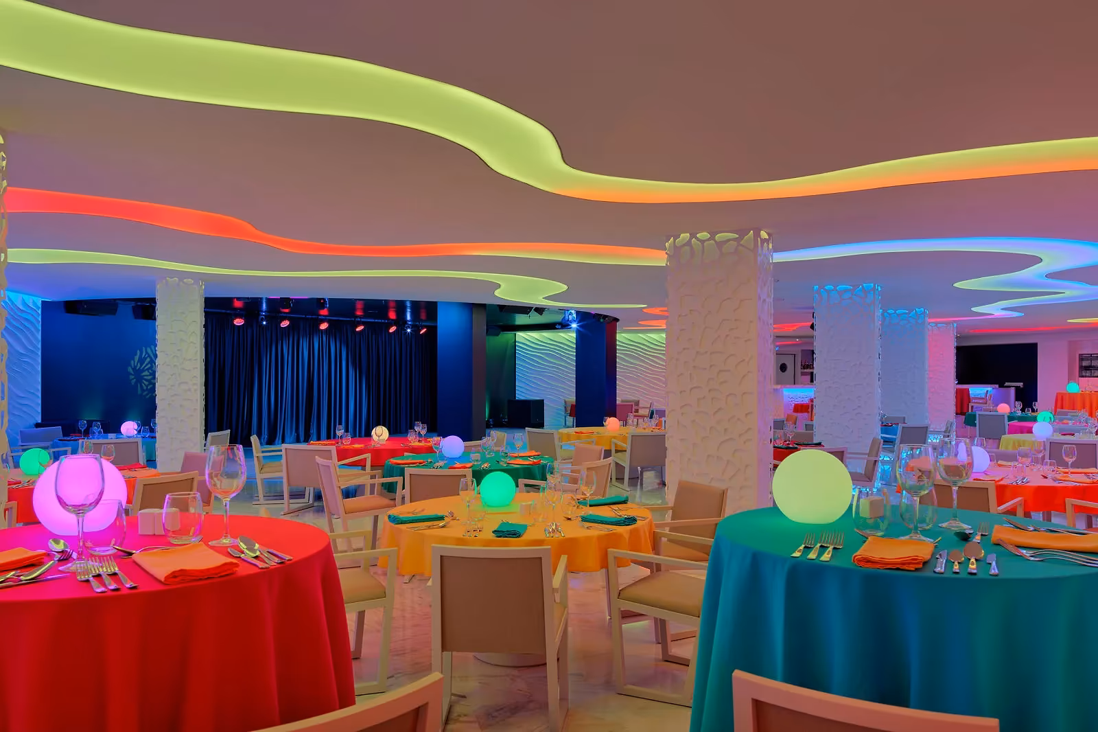 Feestelijk verlichte zaal met ronde tafels in felle kleuren, lichtgevende bollen en een podium met blauw gordijn bij H10 Conquistador in Playa de las Américas