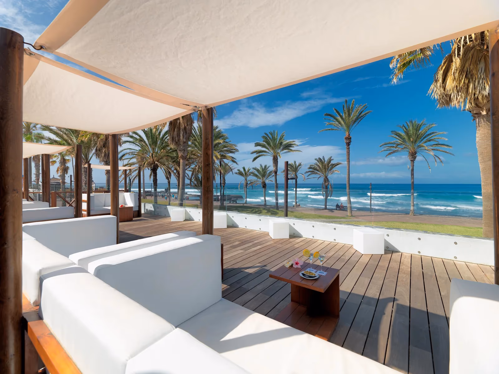 Houten terras met witte loungebanken onder zeildoek, met palmen, de promenade en de zee bij H10 Conquistador in Playa de las Américas