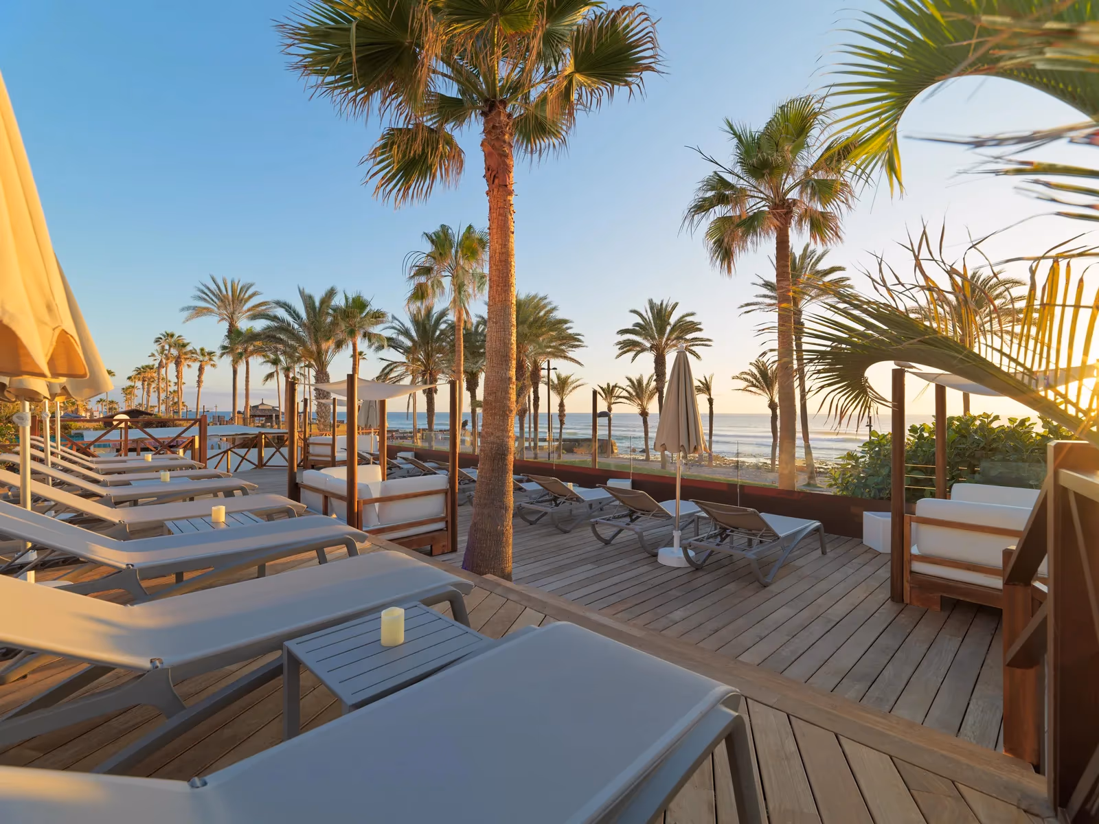 Houten zonneterras met ligbedden en loungebanken in het late zonlicht, met palmen en de oceaan op de achtergrond bij H10 Conquistador in Playa de las Américas