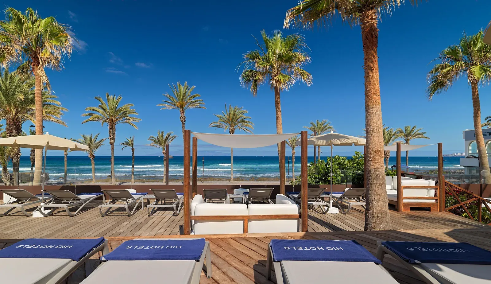 Houten terras met ligbedden en een lounge met witte doeken tegen de oceaan en de palmen bij H10 Conquistador in Playa de las Américas