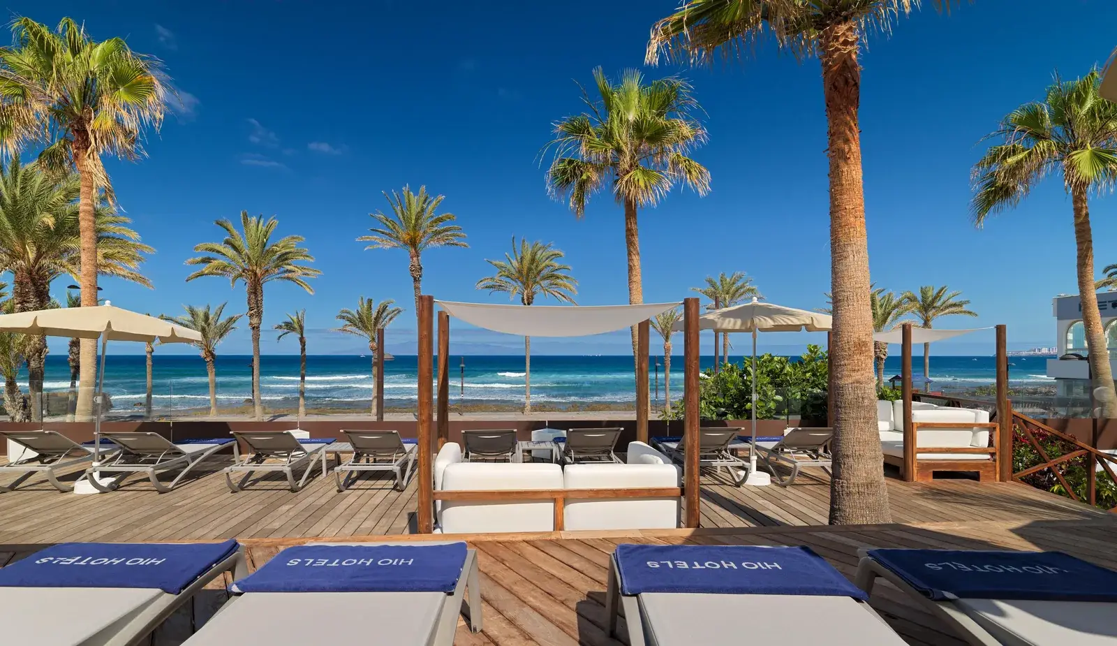 Houten terras met ligbedden en een lounge met witte doeken tegen de oceaan en de palmen bij H10 Conquistador in Playa de las Américas