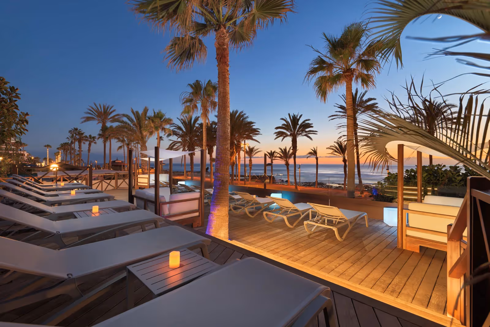 Houten terras met ligbedden en kaarslichtjes in de schemering, met palmen en de zee op de achtergrond bij H10 Conquistador in Playa de las Américas
