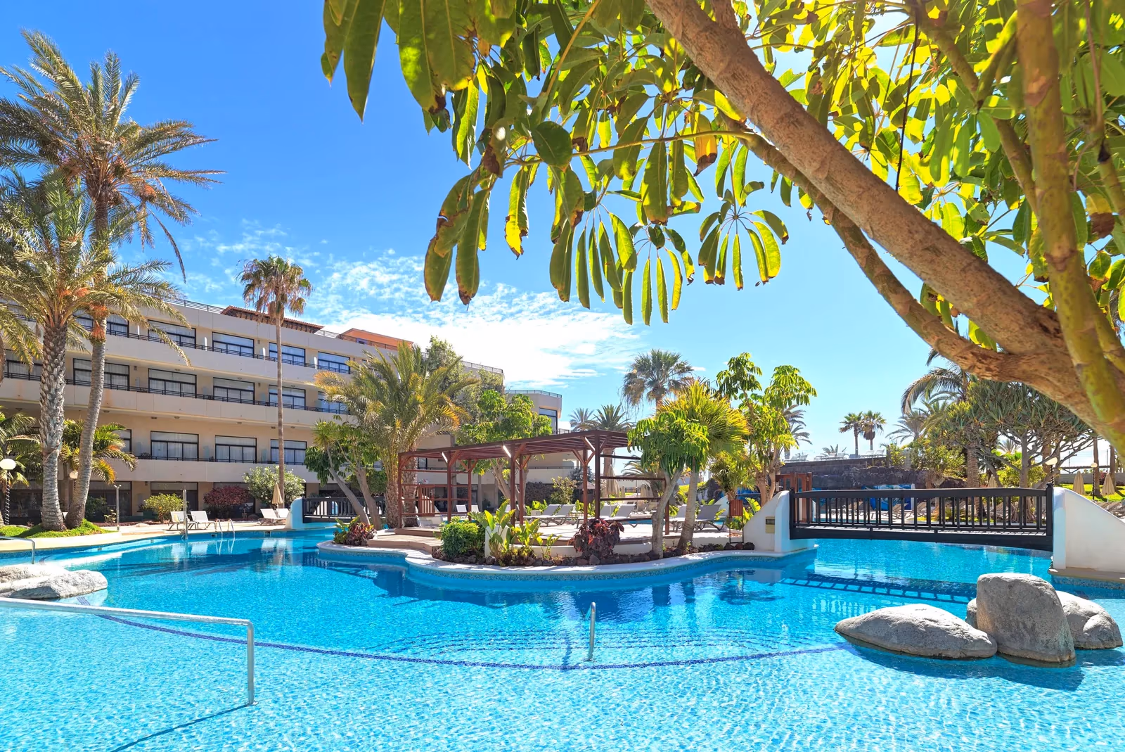 Zwembad met rotsblokken in het water, een houten pergola en een brug tussen palmen, met een hotelvleugel erachter bij H10 Conquistador in Playa de las Américas