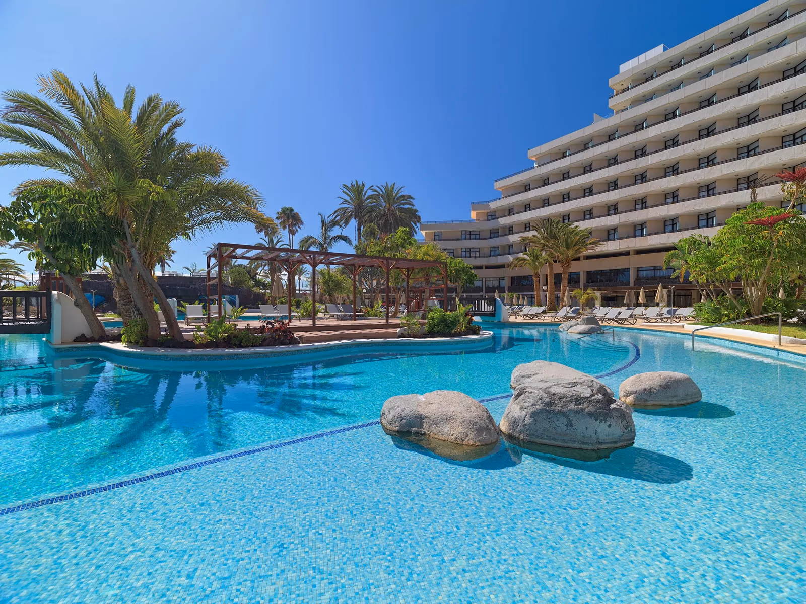 Zwembad met grote rotsblokken en een houten pergola, met een hotelvleugel op de achtergrond bij H10 Conquistador in Playa de las Américas