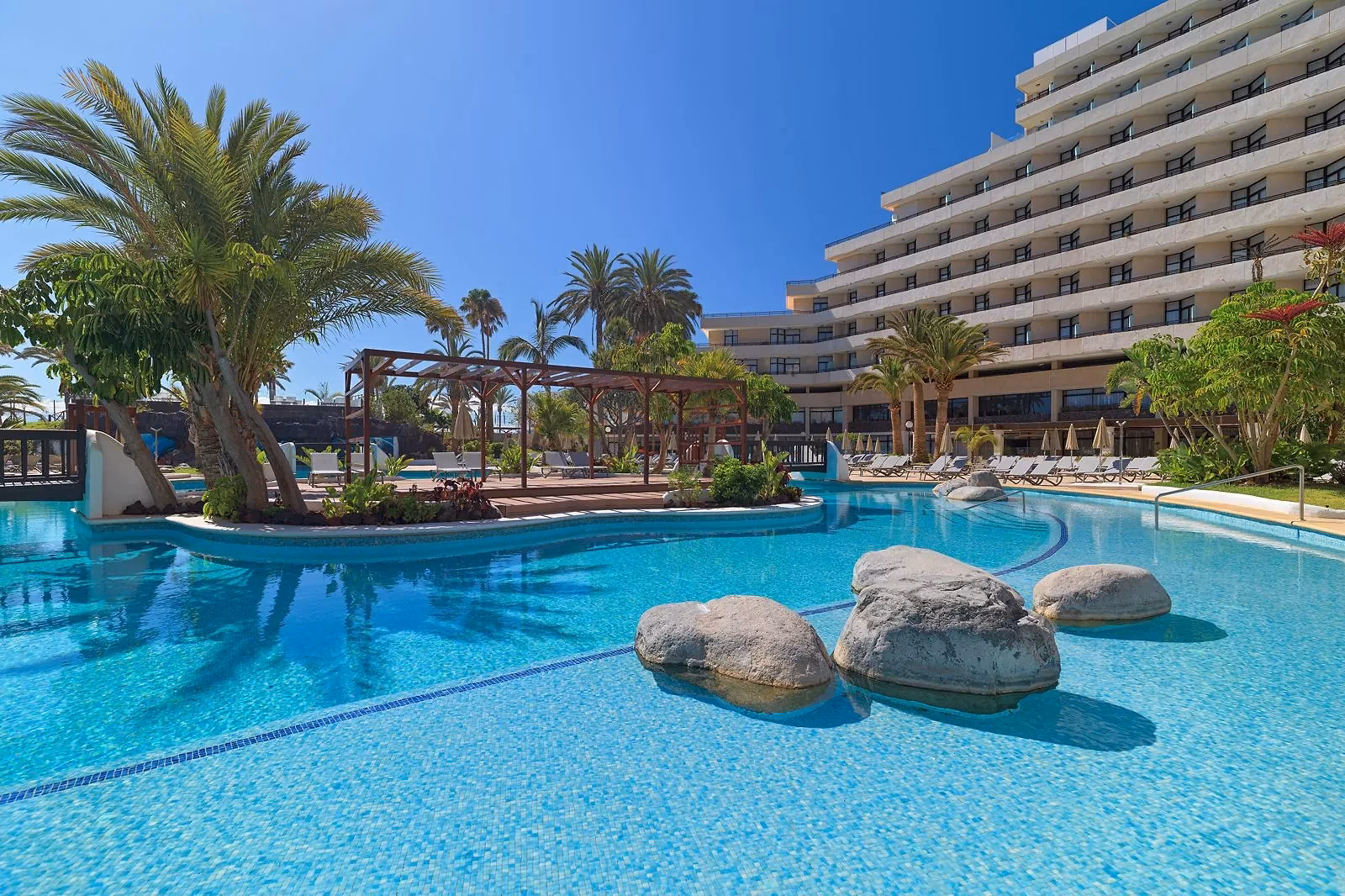 Zwembad met drie rotsblokken en een pergola tussen de palmen, met een grote hotelvleugel rechts bij H10 Conquistador in Playa de las Américas